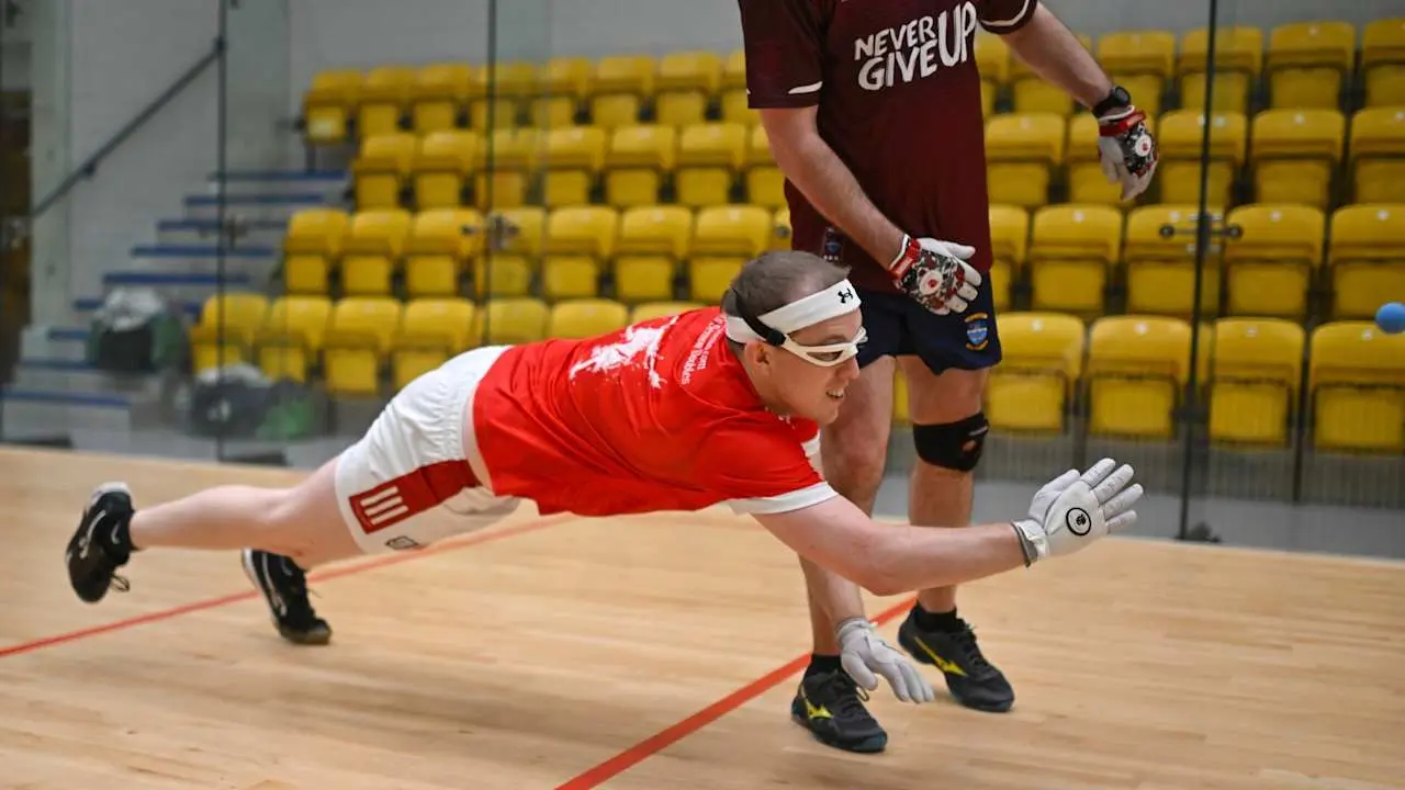 Player in red and white diving horizontally across wooden court, wearing headband and gloves, with opponent in maroon behind 