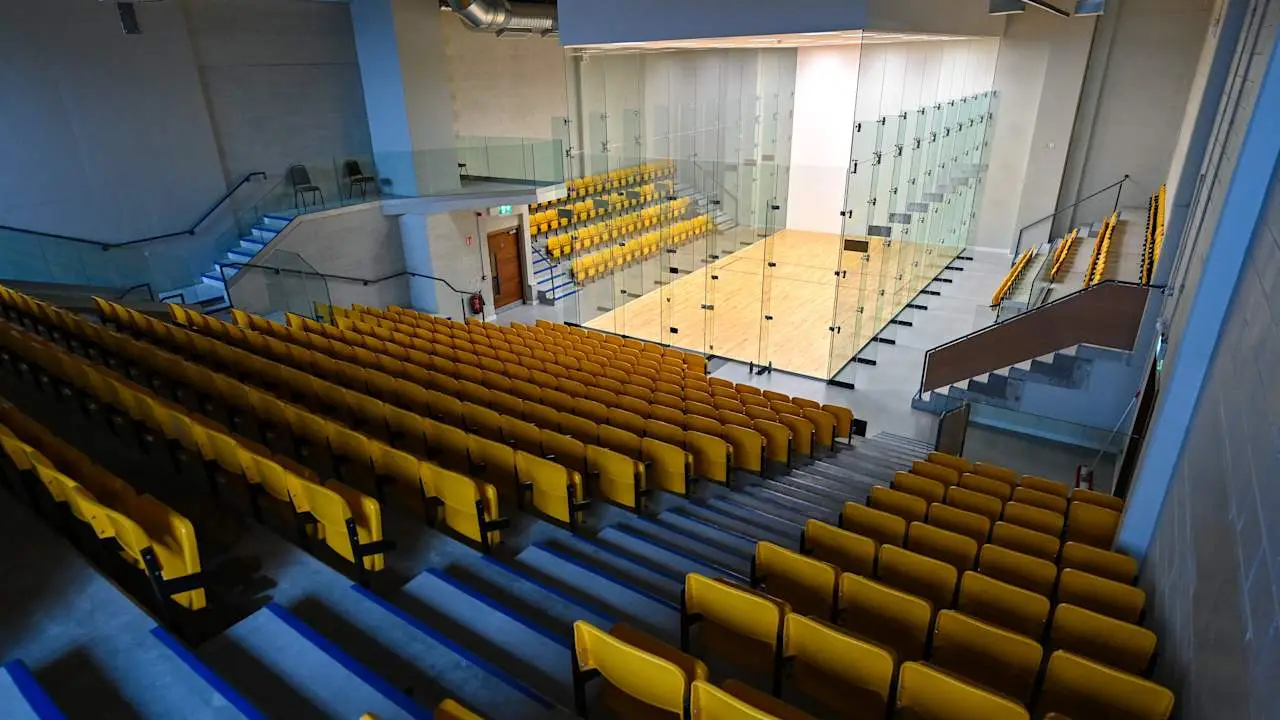 Empty squash court with glass walls surrounded by yellow stadium seating.