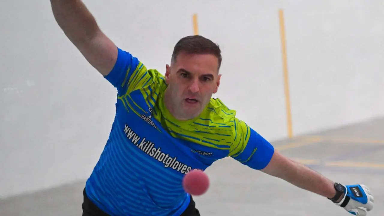 Player in blue and neon yellow Killshot Gloves jersey throwing red handball during match, arm extended mid-throw.