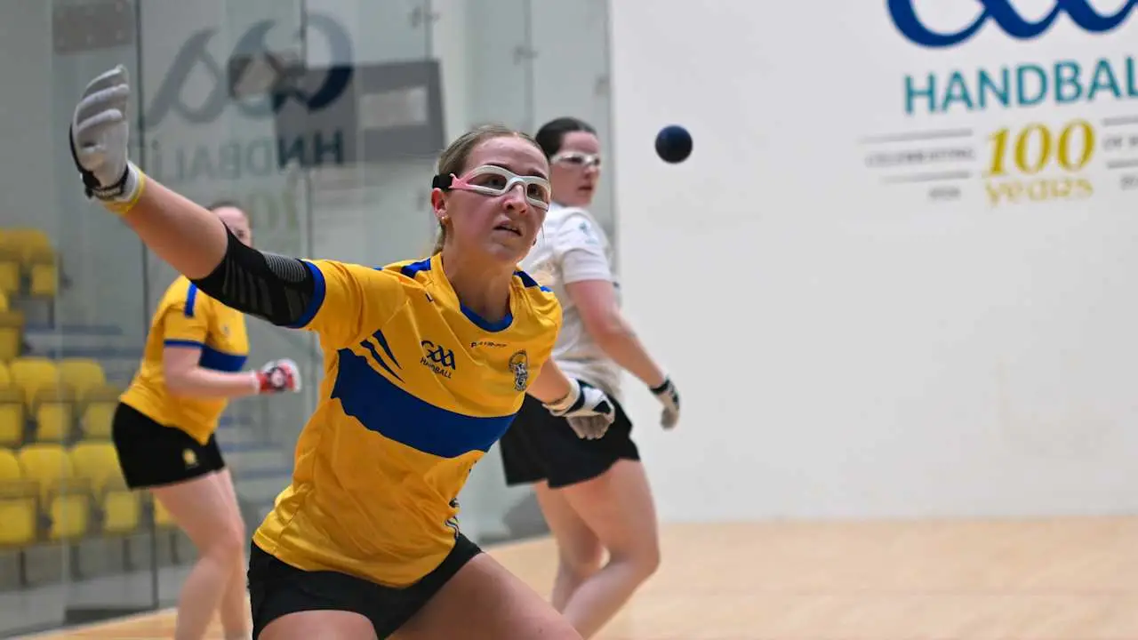 Female GAA handball player in yellow and blue jersey throwing ball on indoor court during match.