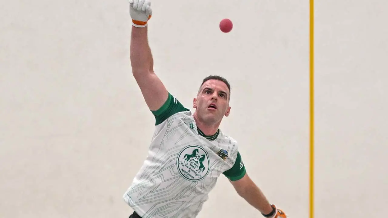 Athlete in white and green GAA Handball kit throwing a red ball overhead on an indoor court, wearing protective gloves and wr