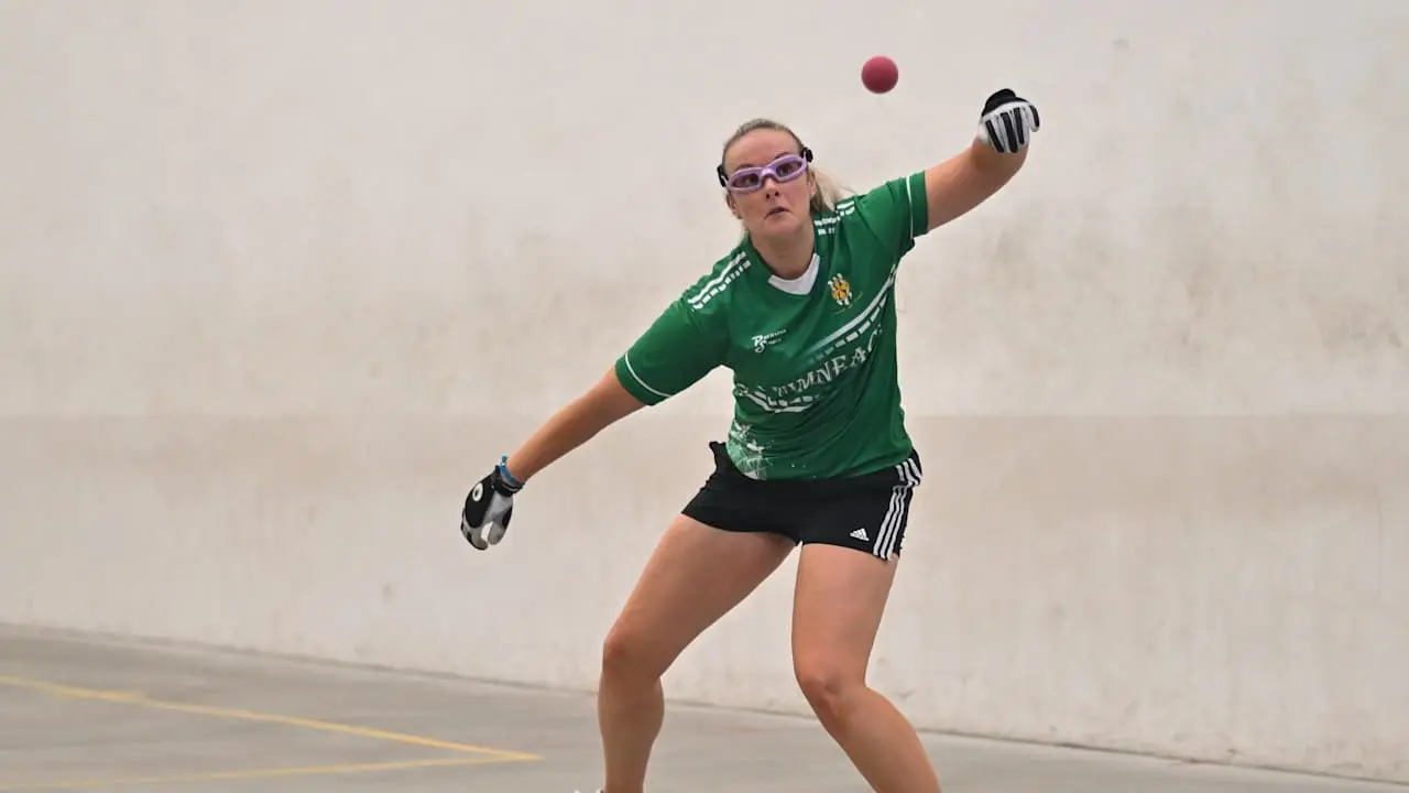 Female handball player in green jersey reaching to catch red ball on indoor court, wearing purple glasses and black gloves.