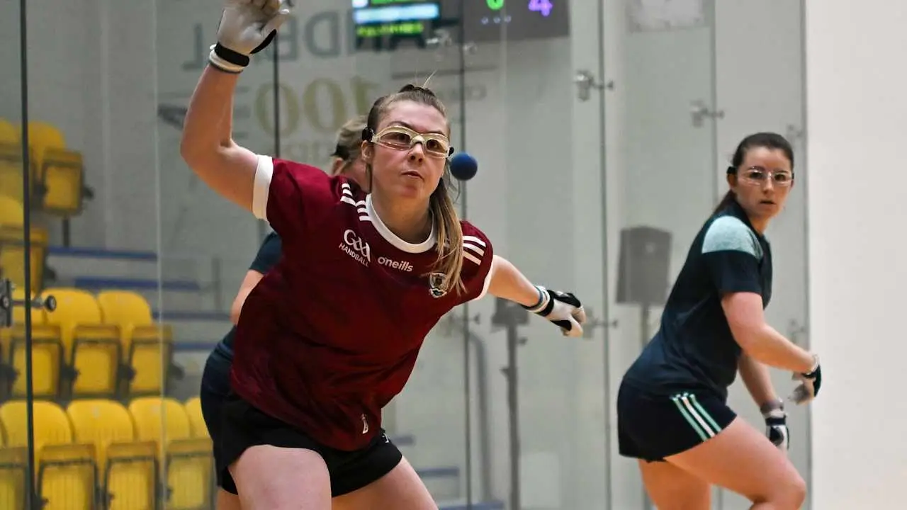 Young player in maroon and navy GAA handball jersey throws blue ball during match on indoor court.