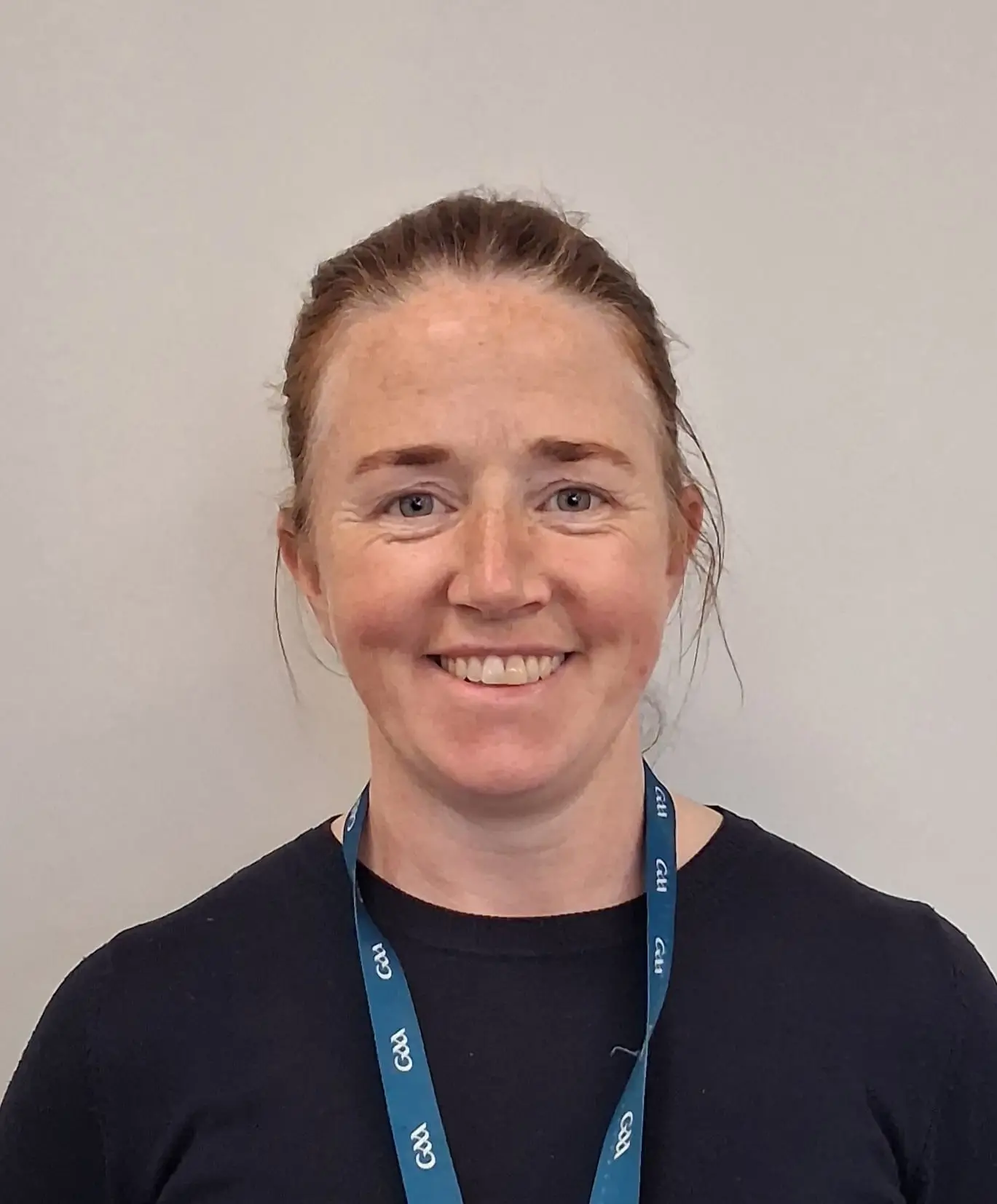 Smiling person with brown hair, wearing a dark shirt and a blue lanyard, standing against a plain background.