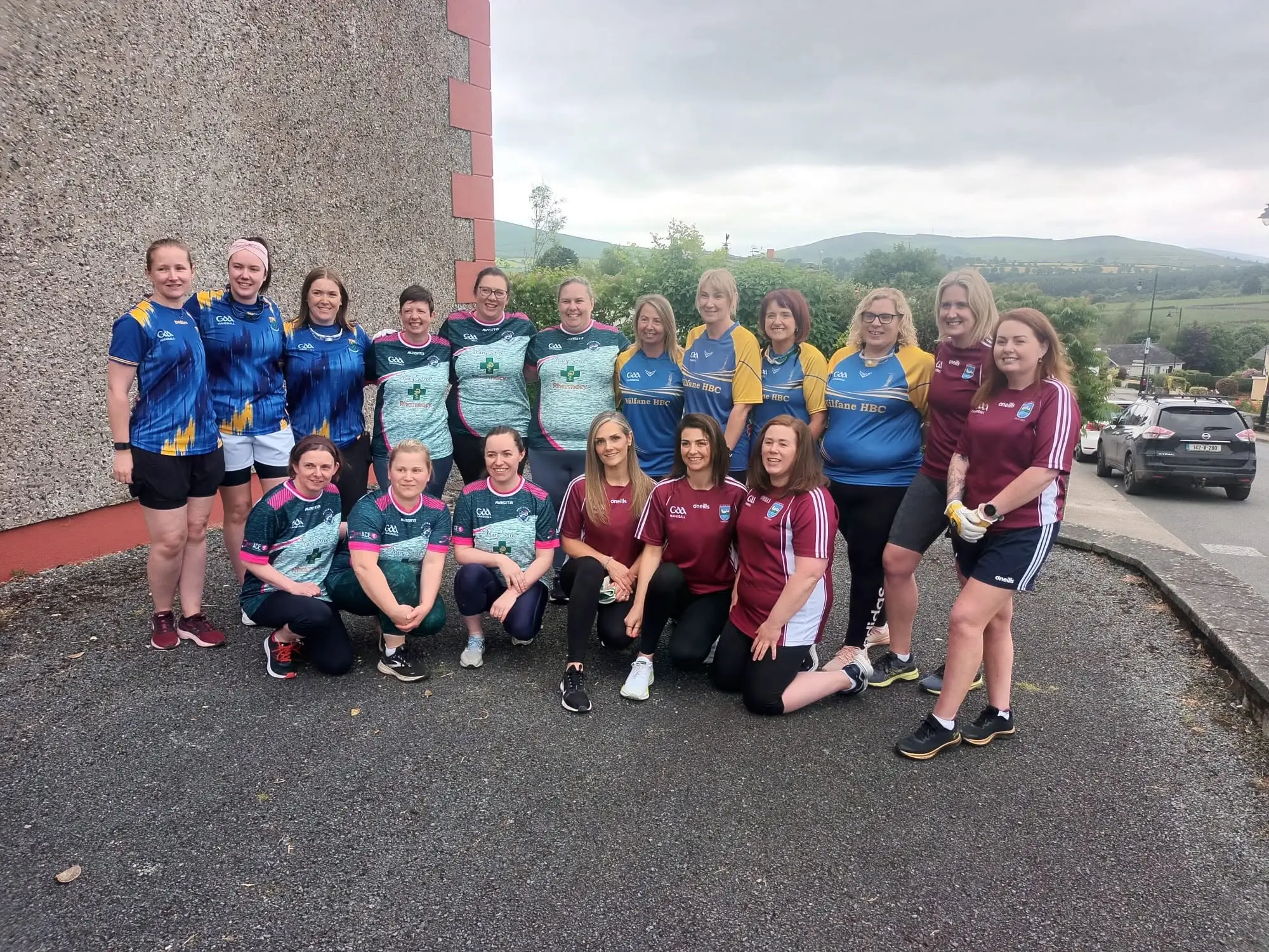 Two women's GAA handball teams pose together outdoors against a building, with rural Irish landscape in background.