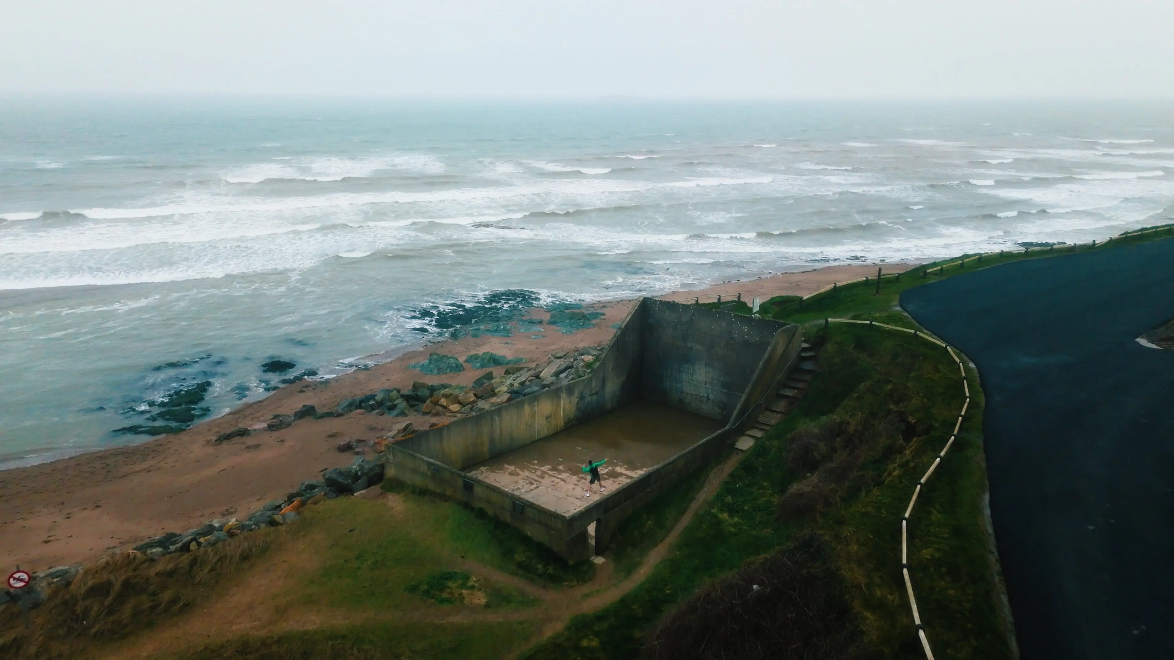 Aerial view of a coastal area with crashing waves, rocky beach, and a person in a concrete structure near a curved road and green grassy patches.