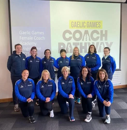 Twelve female gaelic games coaches in blue and navy tracksuits pose in two rows in front of a screen displaying "Gaelic Games