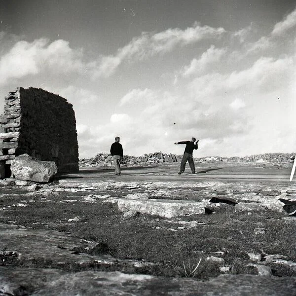 Two people stand near stone ruins on a rocky landscape under a cloudy sky. One person swings a golf club.