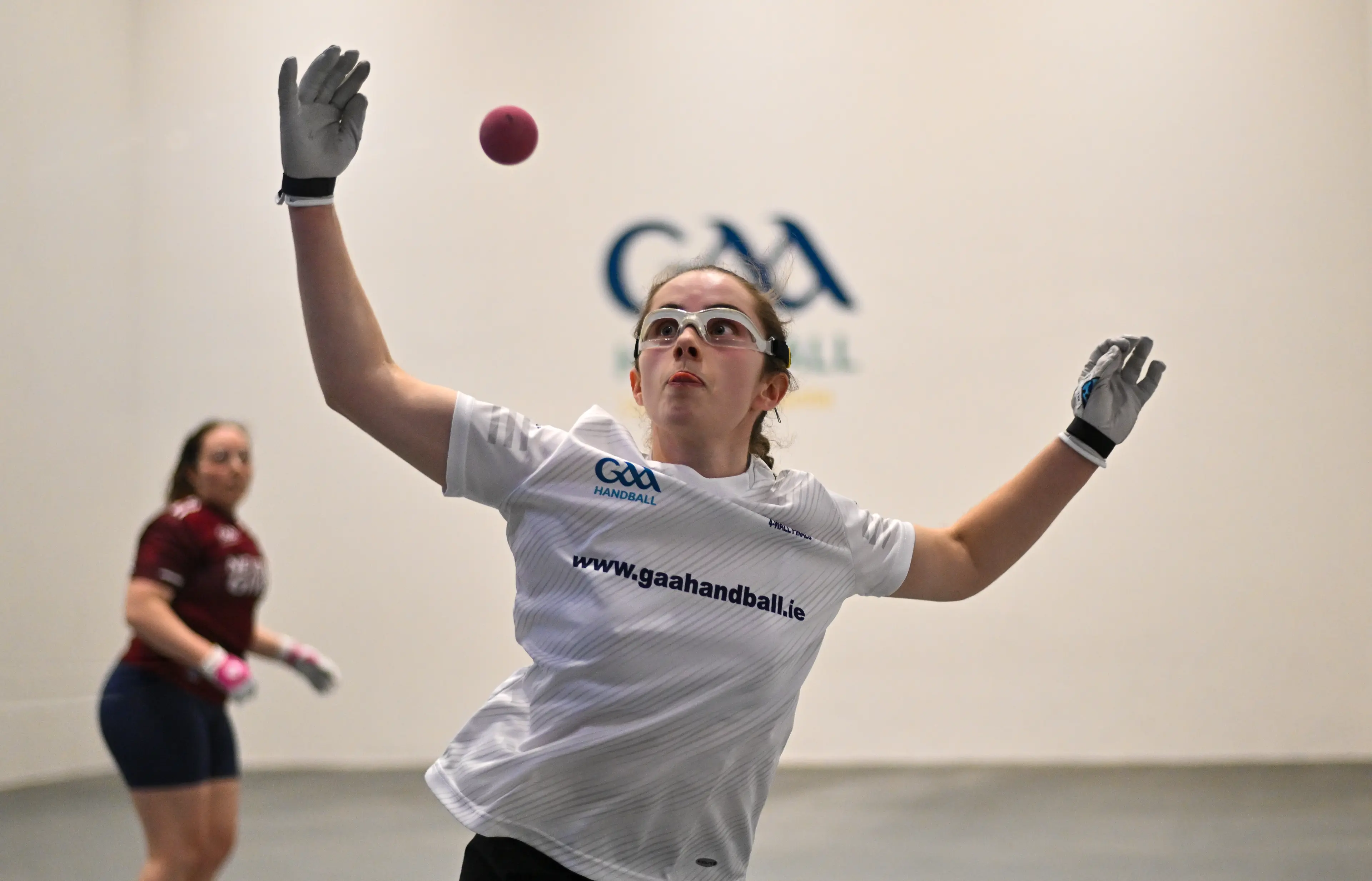Female GAA handball player in white jersey jumping to catch maroon ball on indoor court, opponent in maroon visible in backgr