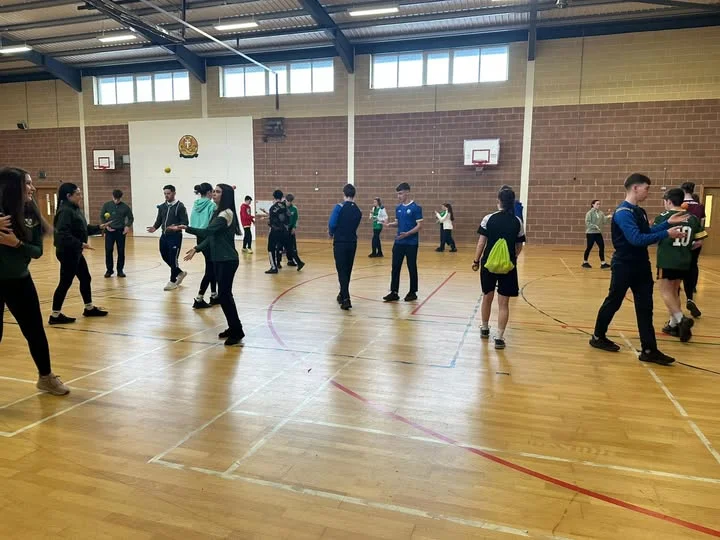 Students participating in a PE class or training session in a school gymnasium with wooden floors and brick walls.